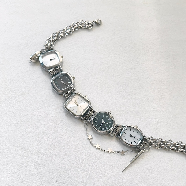 May include: A silver bracelet with five square-shaped watches attached. The watches have silver bands and black or white faces. A silver chain with star charms hangs from the bracelet.