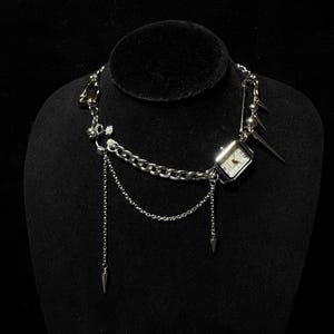 May include: A silver chain necklace with a small square watch, safety pin, and spikes. The necklace has multiple chains and dangles.