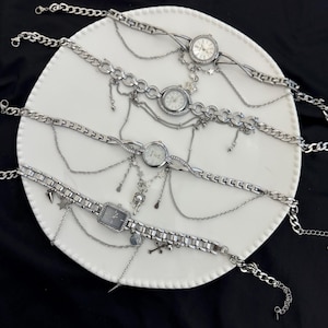 Puede incluir: Cuatro pulseras de reloj plateadas con cadenas y abalorios decorativos se exhiben sobre un plato blanco sobre un fondo negro. Los relojes tienen esferas redondas y rectangulares, con varios adornos.