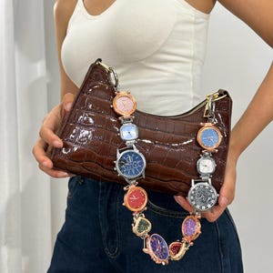 May include: A brown crocodile-embossed handbag with a chain strap made of linked watches with colorful faces. The bag is being held by a person wearing blue jeans.
