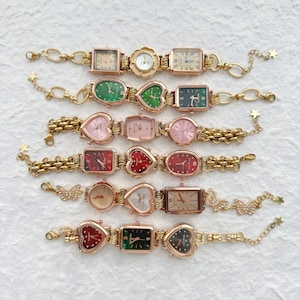 May include: A set of nine gold-tone wristwatches with different coloured faces and designs. The watches have heart, oval, and square-shaped faces. The watches are on gold-tone chains with decorative charms.
