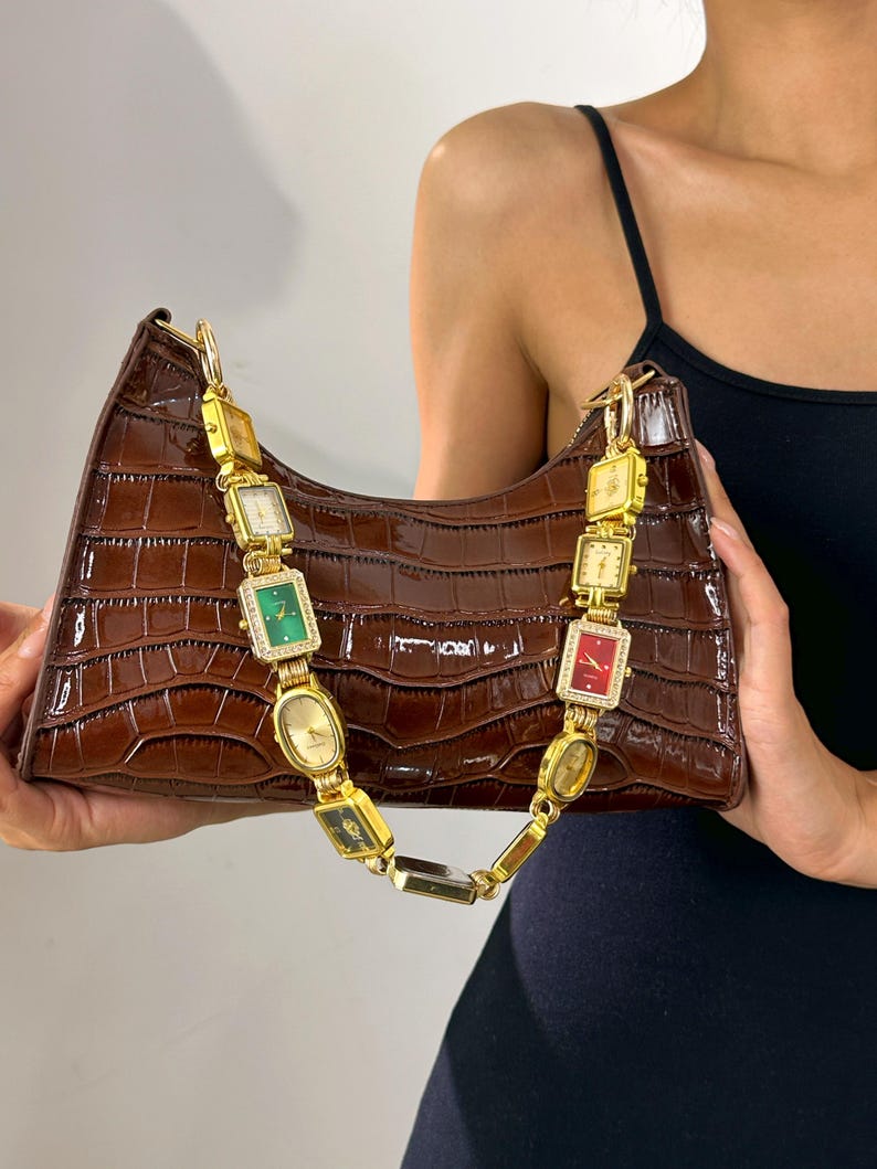 May include: A brown, crocodile-textured handbag with a gold chain strap made of vintage watches. The watches have various colored faces and are connected by gold links. The bag is held up by a person.