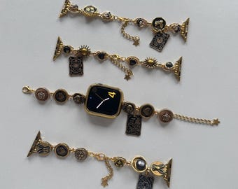 Könnte beinhalten: Goldfarbene Apple Watch-Armbänder mit dekorativen Anhängern. Die Armbänder verfügen über verschiedene Anhänger, darunter Sonnen, Monde und Anhänger im Tarotkarten-Stil. Ein Armband ist an einer goldenen Apple Watch befestigt, die die Uhrzeit anzeigt.