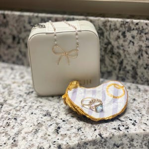 May include: A white jewelry box with a gold bow and the word "KJH" on the front. A white and purple striped oyster shell ring dish with gold accents and a ring with a blue stone.