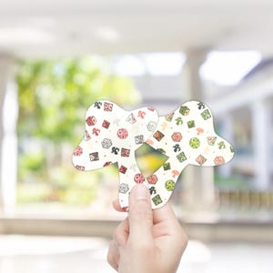 Könnte beinhalten: Zwei cremefarbene, würfelförmige Spielsteine mit einem Muster aus Würfeln, Pilzen und anderen spielbezogenen Grafiken. Die Teile werden vor einem verschwommenen Hintergrund gehalten, der das detaillierte Design und die Farben zeigt.