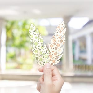 Könnte beinhalten: Ein dekorativer, federförmiger Ausschnitt mit einem detaillierten Muster aus kleinen, skurrilen Illustrationen in Grün-, Orange- und Brauntönen. Die Feder wird vor einem verschwommenen Hintergrund gehalten, der ihr aufwendiges Design zeigt.