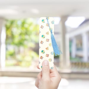 Puede incluir: Un marcapáginas crema con ilustraciones de dados coloridos y una borla azul. El marcapáginas, sostenido en una mano, muestra una forma rectangular y diseños detallados de dados. La borla añade un elemento decorativo.