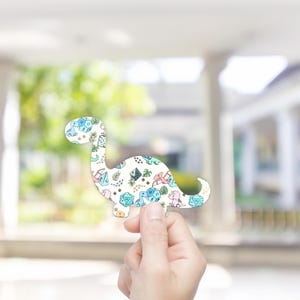 Puede incluir: Un recorte blanco con forma de dinosaurio con un patrón colorido de dados, dinosaurios y follaje. El diseño presenta tonos de azul, rosa y verde. El recorte está sostenido por una mano.