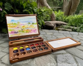 May include: A wooden watercolor paint set with a landscape painting in progress. The set includes a palette of various colors, a brush, and a lid. The painting features a colorful field with blue sky.