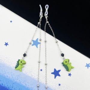 Peut inclure: Deux chaînes de lunettes argentées avec des breloques de dinosaures verts et des perles noires. Les chaînes sont attachées à des boucles en silicone transparentes.