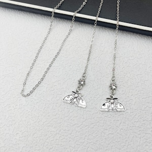 Op de afbeelding: Zilverkleurige ketting met een fijne ketting en een mot-hanger. De mot-hanger heeft ingewikkelde details en hangt onder een kleine, sprankelende edelsteen. De ketting wordt getoond op een lichtgekleurd oppervlak.