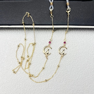 Könnte beinhalten: Goldfarbene Brillenketten mit filigranem Design. Jede Kette hat kleine goldene Perlen und einen Anhänger mit einer Katzensilhouette, die auf einem Mond sitzt. Die Ketten haben auch kleine rote Edelsteine.