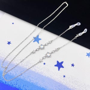 Op de afbeelding: Twee zilveren brillenkettingen met heldere kristallen accenten. Elke ketting heeft een grotere kristal in het midden en kleinere kristallen langs de ketting. De kettingen zijn bevestigd aan heldere siliconen lussen.