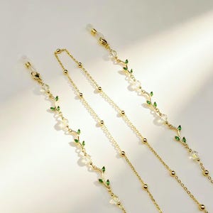 May include: A gold-toned glasses chain featuring delicate vine-like links adorned with small green enamel leaves and clear crystal beads. The chain includes two clear silicone loops at the ends for attachment to eyewear frames, with a thin metal link chain.