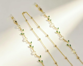 May include: A gold-toned glasses chain featuring delicate vine-like links adorned with small green enamel leaves and clear crystal beads. The chain includes two clear silicone loops at the ends for attachment to eyewear frames, with a thin metal link chain.