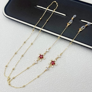 May include: Gold-tone glasses chain with a delicate design. The chain features small, round beads and star-shaped accents with red and white gemstones. The chain is designed to hold spectacles.