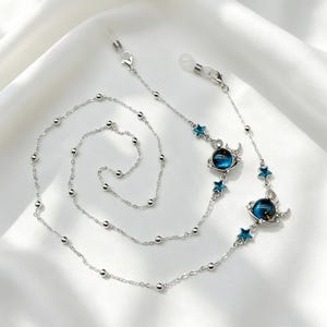 Peut inclure: Une chaîne de lunettes argentée composée de petites perles rondes sur une fine chaîne à maillons. Le design comprend deux breloques en forme de planète bleue, entourées d'un anneau argenté et d'un croissant de lune, avec des étoiles bleues.