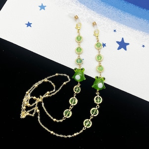 May include: A gold chain with green frog charms and green crystal beads. The chain is designed to hold glasses.