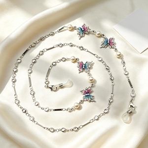 Op de afbeelding: Een zilverkleurige brillenketting met kleine witte parels en vlindervormige bedels met blauwe en roze emaille details. De ketting is opgebouwd uit afwisselende ronde en rechthoekige schakels en is voorzien van doorzichtige siliconen lussen.