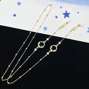 Könnte beinhalten: Zwei goldene Brillenketten mit bunten Strasssteinen. Jede Kette hat einen runden Anhänger mit einem Blumenmuster.