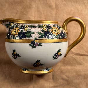 May include: A white ceramic creamer with a gold handle and trim. The creamer is decorated with yellow, blue, and green floral patterns. The handle and trim are painted gold.