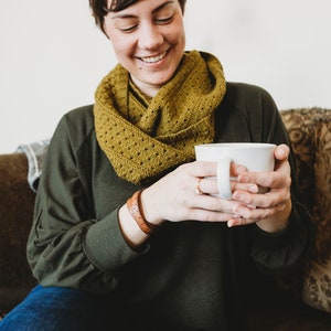 May include: A woman wearing a green sweater and a yellow knitted scarf sits on a couch and holds a white mug in both hands.