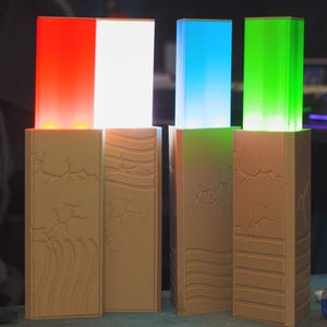 May include: Four illuminated rectangular light fixtures with textured, tan bases. The lights glow red, white, blue, and green. The bases have carved designs of cracks and waves. The lights are likely decorative or for ambiance.