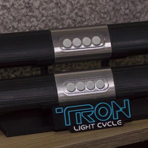 Può includere: Due oggetti di scena neri e argentati della light cycle del film Tron. Ogni oggetto di scena ha un'impugnatura nera con dettagli argentati e quattro pulsanti. Gli oggetti di scena sono esposti su un supporto nero con il logo Tron Light Cycle.