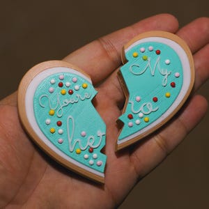 May include: Two turquoise heart-shaped cookies with white icing and colorful sprinkles. One cookie says "You're my" and the other says "hero".