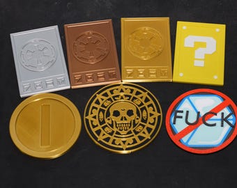 Puede incluir: Una colección de seis fichas y tarjetas decorativas que incluye tres tarjetas rectangulares con símbolos imperiales, una tarjeta de bloque de interrogación amarilla, una moneda dorada, un medallón de calavera azteca dorado y un letrero circular con el texto FUCK.