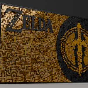 May include: A black and gold box featuring the word "ZELDA" in black lettering. The box has a repeating spiral pattern on one side and a gold emblem with a sword on the other. The box is a collectible item.
