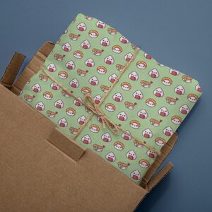 May include: A green fabric with a repeating pattern of cartoon sushi and rice balls, tied with twine, inside a cardboard box. The sushi includes shrimp, and the rice balls have smiling faces. The fabric is a light green color.