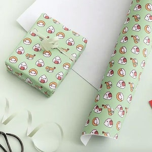May include: Gift wrap featuring a repeating pattern of cute sushi and rice ball illustrations on a light green background. A wrapped gift with a white ribbon and a roll of matching paper are visible.