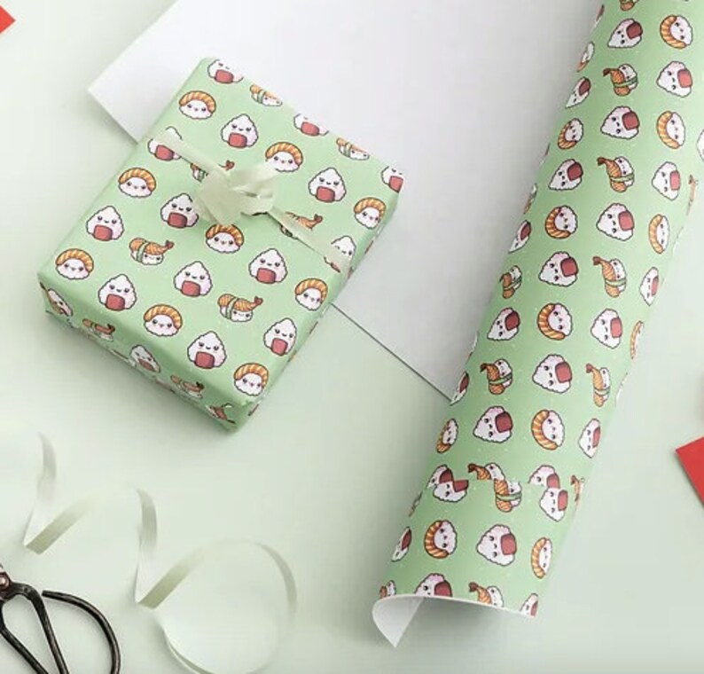 May include: Gift wrap featuring a repeating pattern of cute sushi and rice ball illustrations on a light green background. A wrapped gift with a white ribbon and a roll of matching paper are visible.