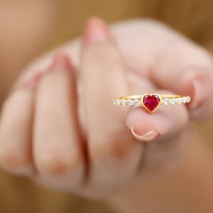 May include: A gold ring featuring a heart-shaped red gemstone at the center, surrounded by small, clear gemstones. The ring is held up, showcasing its design and sparkle. The ring is a piece of jewelry.