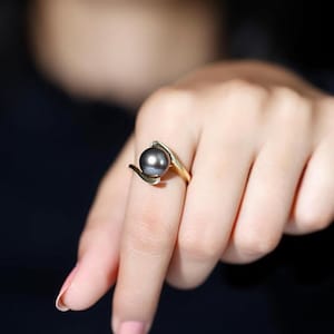 May include: A gold ring featuring a dark gray pearl. The ring's design has a flowing, organic shape that cradles the pearl. The pearl is round and smooth, and the ring is set on a finger.
