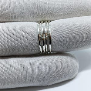 Puede incluir: Un anillo de plata con un dise&ntilde;o &uacute;nico. El anillo presenta m&uacute;ltiples bandas unidas por un peque&ntilde;o conector circular. Las bandas son lisas y brillantes, reflejando la luz. El anillo se muestra sobre un fondo de tela gris.