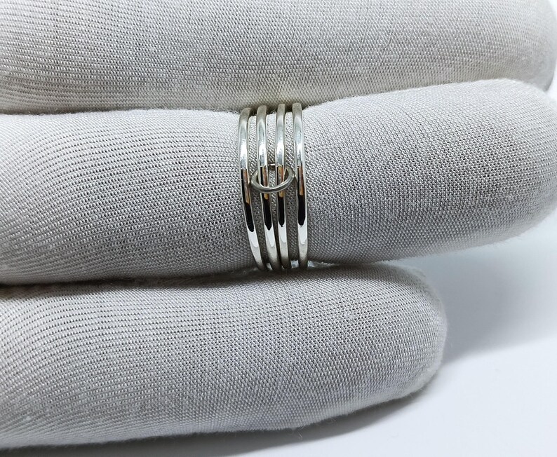 Puede incluir: Un anillo de plata con un dise&ntilde;o &uacute;nico. El anillo presenta m&uacute;ltiples bandas unidas por un peque&ntilde;o conector circular. Las bandas son lisas y brillantes, reflejando la luz. El anillo se muestra sobre un fondo de tela gris.