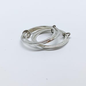 Puede incluir: Un anillo plateado con tres bandas entrelazadas. Las bandas son lisas y brillantes, creando un dise&ntilde;o moderno y minimalista. El anillo se presenta sobre un fondo blanco liso, resaltando su elegante sencillez.