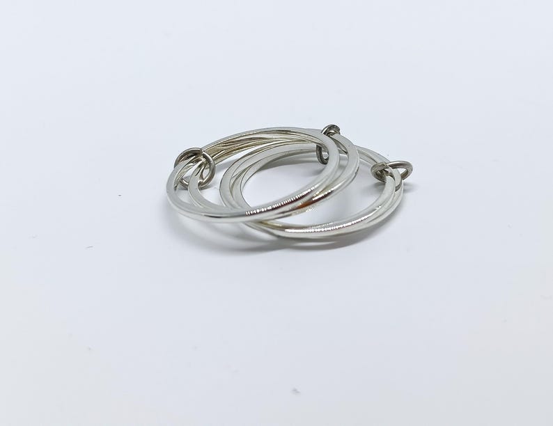Puede incluir: Un anillo plateado con tres bandas entrelazadas. Las bandas son lisas y brillantes, creando un dise&ntilde;o moderno y minimalista. El anillo se presenta sobre un fondo blanco liso, resaltando su elegante sencillez.