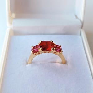 May include: A gold ring featuring three gemstones: a central, large, red square stone, and two smaller, pink rectangular stones. The ring is presented in a white box, suitable for jewellery.