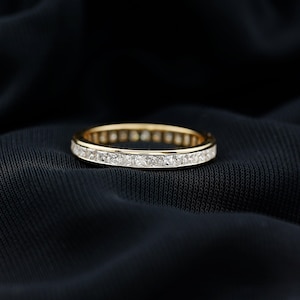 Peut inclure: Une bague en or avec une rangée continue de pierres précieuses claires, taillées en carré. La bague est posée sur un fond sombre et texturé, mettant en valeur l'éclat des pierres et la tonalité chaude de l'or.