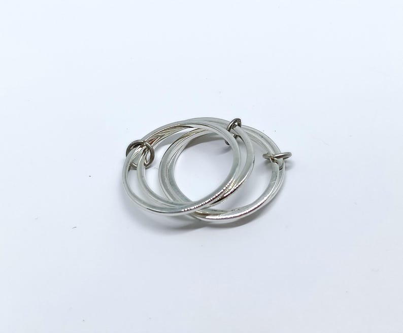 Puede incluir: Un colgante de anillos entrelazados de color plateado. El colgante consta de tres anillos circulares conectados. Los anillos son lisos y brillantes, con un dise&ntilde;o sencillo y elegante. El colgante se muestra sobre un fondo blanco.