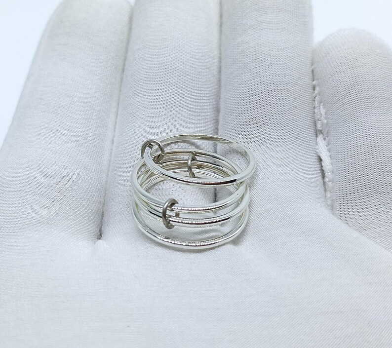 Puede incluir: Un anillo plateado con m&uacute;ltiples bandas conectadas por peque&ntilde;os aros. El anillo, hecho de metal brillante, descansa sobre una superficie blanca. El dise&ntilde;o es minimalista y moderno, adecuado para el uso diario.