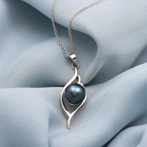 Può includere: Una collana con pendente in argento con una perla grigio scuro. Il pendente ha un design stilizzato e aperto che tiene la perla. La catena della collana è delicata e argentata, su uno sfondo di tessuto azzurro.