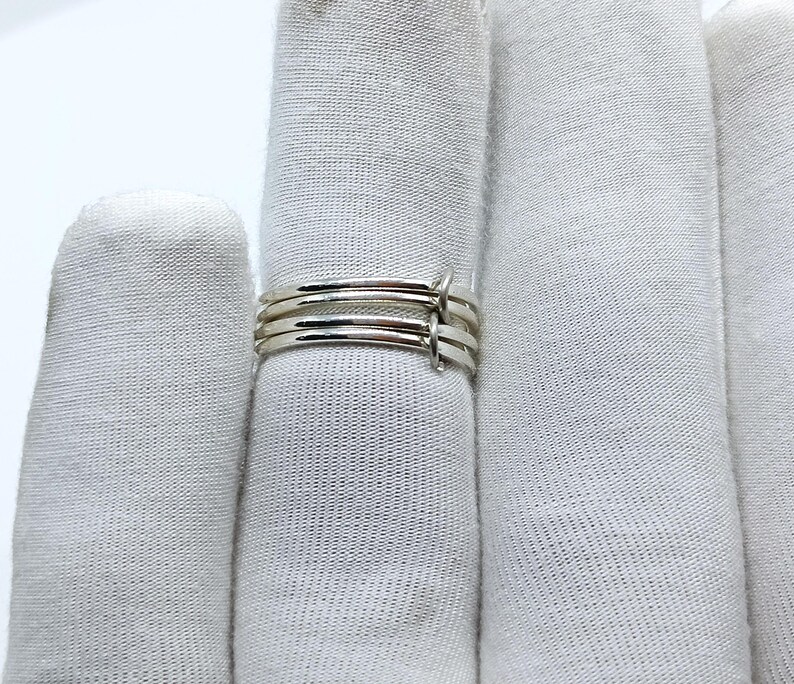 Puede incluir: Un anillo plateado con un dise&ntilde;o minimalista. El anillo presenta cuatro bandas finas unidas por peque&ntilde;os conectores verticales. Las bandas son lisas y brillantes, reflejando la luz. El anillo se muestra sobre un fondo blanco.