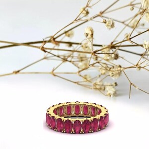 May include: A gold band ring with a row of oval-shaped ruby gemstones. The ring is set on a white background.
