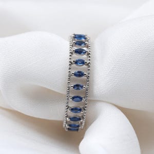 May include: A silver-toned ring featuring a row of marquise-cut blue stones set between two beaded metal bands. The design consists of multiple small, elongated blue gems arranged vertically within a textured, metallic frame structure.