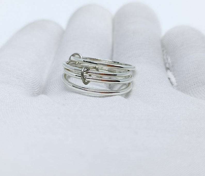 Puede incluir: Un anillo de plata con un dise&ntilde;o &uacute;nico. El anillo presenta m&uacute;ltiples bandas finas conectadas por peque&ntilde;os eslabones curvos. Las bandas son lisas y brillantes, reflejando la luz. El anillo est&aacute; sobre un fondo blanco.