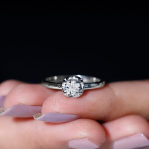 May include: A silver ring with a square-shaped diamond cluster. The ring is set on a finger with light purple nail polish, against a black background. The ring's band is thin and polished.